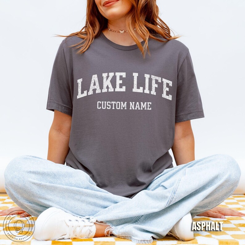 Peut inclure: T-shirt gris asphalte avec le texte "LAKE LIFE" en lettres blanches. En dessous, le texte "CUSTOM NAME". Le mod&egrave;le porte un jean bleu clair et des baskets blanches. Le t-shirt est de "The Quiet Bookish Boutique".
