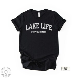 Peut inclure: T-shirt noir avec l'inscription "LAKE LIFE" au-dessus de "CUSTOM NAME" en lettres blanches et us&eacute;es. Le t-shirt a des manches retrouss&eacute;es et un col rond. Le mot "BLACK" est dans le coin inf&eacute;rieur droit.