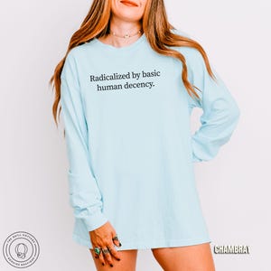 May include: A light blue long-sleeve shirt with the text "Radicalized by basic human decency." The shirt is a solid color and has a relaxed fit. The image also includes the text "CHAMBRAY" and a logo for "The Quill Cottage Bookish Boutique."