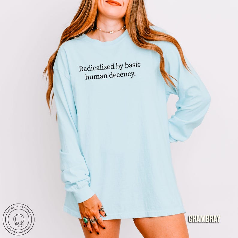 May include: A light blue long-sleeve shirt with the text "Radicalized by basic human decency." The shirt is a solid color and has a relaxed fit. The image also includes the text "CHAMBRAY" and a logo for "The Quill Cottage Bookish Boutique."