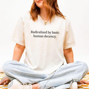 May include: Ivory-colored t-shirt with the text "Radicalized by basic human decency." The shirt is a short-sleeved, crew neck style. The model is wearing light blue jeans and sitting on a yellow and white checkered surface.