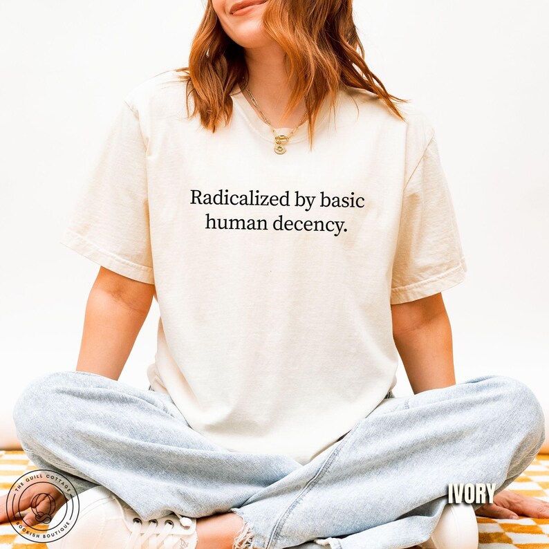 May include: Ivory-colored t-shirt with the text "Radicalized by basic human decency." The shirt is a short-sleeved, crew neck style. The model is wearing light blue jeans and sitting on a yellow and white checkered surface.