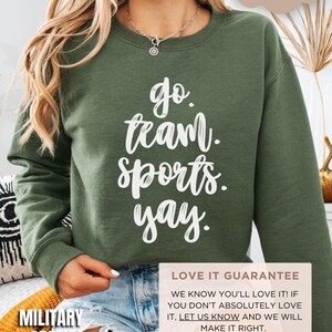 May include: A green sweatshirt with the text 'go team sports yay.' printed in white. The sweatshirt is displayed on a model wearing denim shorts. The text 'COZY SOFT' is in a pink cloud shape in the upper right corner of the image. The text 'MILITARY GREEN' is in the lower left corner of the image. The text 'LOVE IT GUARANTEE' is in a pink rectangle in the lower right corner of the image. The text 'WE KNOW YOU'LL LOVE IT! IF YOU DON'T ABSOLUTELY LOVE IT, LET US KNOW AND WE WILL MAKE IT RIGHT. We're only a message away!' is in the pink rectangle below the text 'LOVE IT GUARANTEE'.