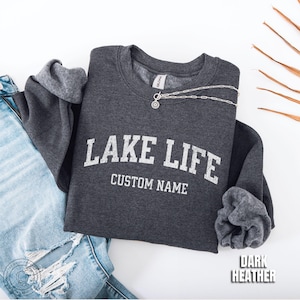 May include: A dark heather gray crewneck sweatshirt featuring the white text LAKE LIFE and CUSTOM NAME in a collegiate font. The garment has long sleeves with ribbed cuffs and a Gildan brand label at the inner neckline. A silver chain necklace rests on the collar.