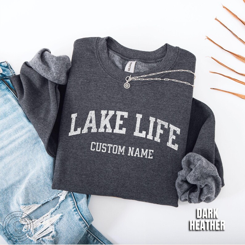 May include: A dark heather gray crewneck sweatshirt featuring the white text LAKE LIFE and CUSTOM NAME in a collegiate font. The garment has long sleeves with ribbed cuffs and a Gildan brand label at the inner neckline. A silver chain necklace rests on the collar.