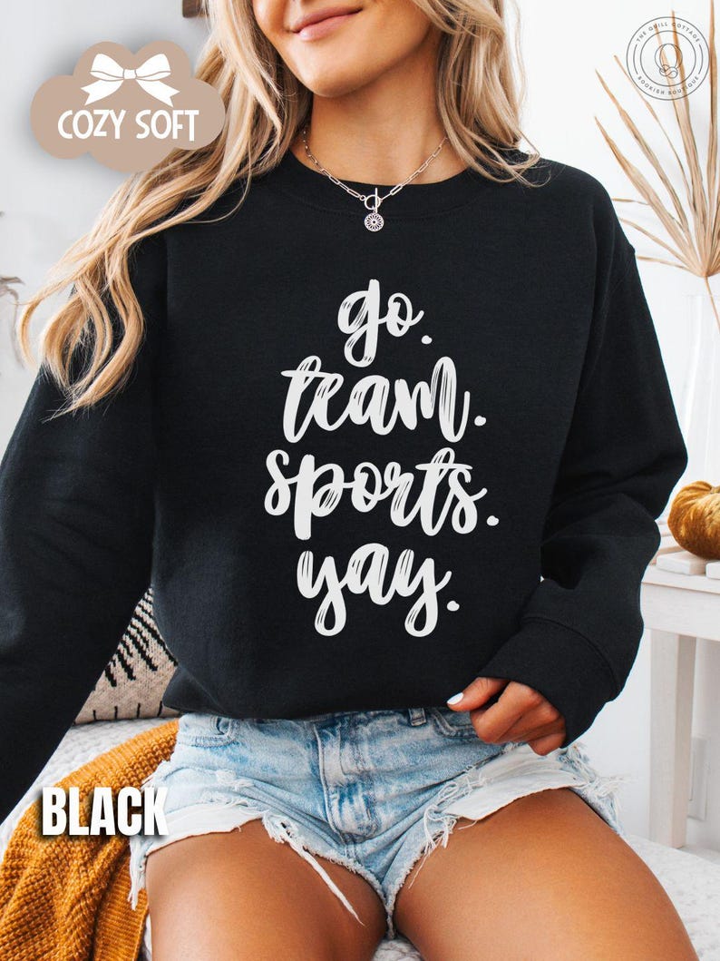 May include: A black sweatshirt with white text that reads 'go team sports yay.' The sweatshirt is being worn by a person wearing denim shorts.