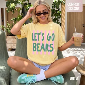 May include: A pale yellow short-sleeve t-shirt featuring the green text LET'S GO BEARS layered over vertical pink stripes. The shirt has a crew neckline and a relaxed fit, with a small Comfort Colors label visible in the upper right corner.