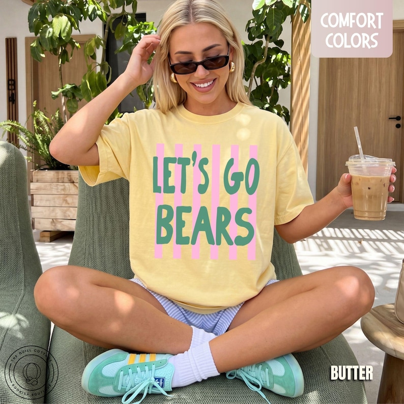 May include: A pale yellow short-sleeve t-shirt featuring the green text LET'S GO BEARS layered over vertical pink stripes. The shirt has a crew neckline and a relaxed fit, with a small Comfort Colors label visible in the upper right corner.