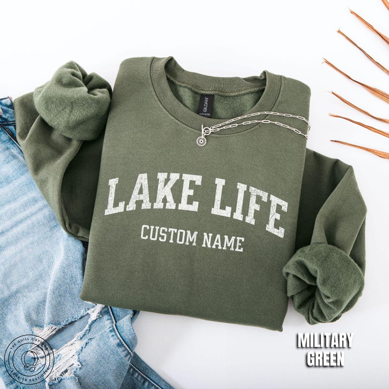 May include: A military green crewneck sweatshirt featuring the white distressed text LAKE LIFE and CUSTOM NAME printed on the front. The garment is paired with blue distressed denim jeans and a silver-toned chain necklace with a circular pendant.