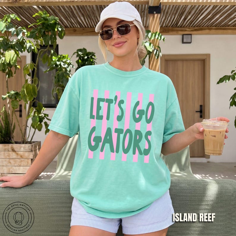 May include: A mint green short-sleeve t-shirt featuring vertical pink stripes and the dark green text "LET'S GO GATORS" printed on the front. The shirt is paired with light blue and white pinstriped shorts, a white baseball cap, and sunglasses.