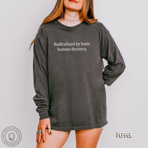 May include: Dark gray long-sleeve shirt with the text "Radicalized by basic human decency." The shirt has a vintage, washed look. The Quill Cottage Bookish Boutique logo is visible in the lower left corner, and the word "Pepper" is in the lower right.