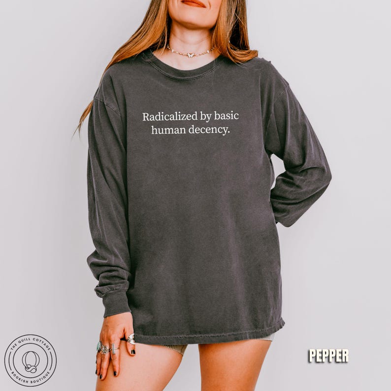 May include: Dark gray long-sleeve shirt with the text "Radicalized by basic human decency." The shirt has a vintage, washed look. The Quill Cottage Bookish Boutique logo is visible in the lower left corner, and the word "Pepper" is in the lower right.