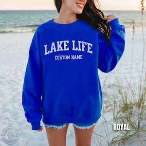 May include: A royal blue long-sleeve crewneck sweatshirt featuring the white distressed text LAKE LIFE arched over the words CUSTOM NAME. The garment has a ribbed collar, cuffs, and hem, and is displayed in a solid, vibrant blue color.