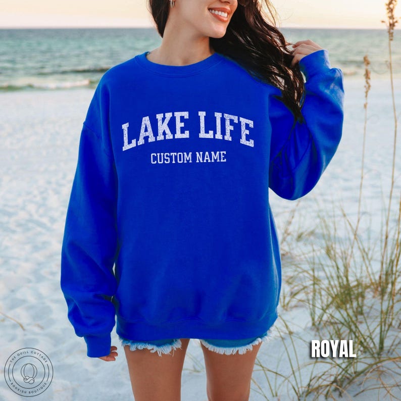 May include: A royal blue long-sleeve crewneck sweatshirt featuring the white distressed text LAKE LIFE arched over the words CUSTOM NAME. The garment has a ribbed collar, cuffs, and hem, and is displayed in a solid, vibrant blue color.