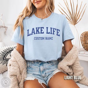 Peut inclure: T-shirt bleu clair avec l'inscription "LAKE LIFE" en lettres majuscules bleu marine au-dessus de "CUSTOM NAME". Le t-shirt est &agrave; manches courtes et port&eacute; avec un short en jean. Les mots "LIGHT BLUE" sont dans un encadr&eacute; blanc en bas &agrave; droite.