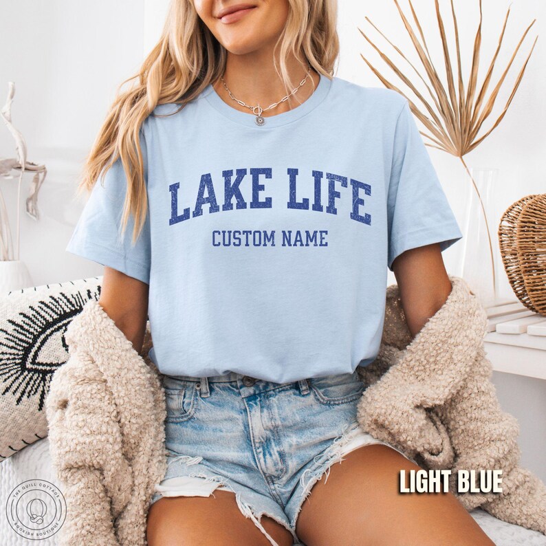 Peut inclure: T-shirt bleu clair avec l'inscription "LAKE LIFE" en lettres majuscules bleu marine au-dessus de "CUSTOM NAME". Le t-shirt est &agrave; manches courtes et port&eacute; avec un short en jean. Les mots "LIGHT BLUE" sont dans un encadr&eacute; blanc en bas &agrave; droite.