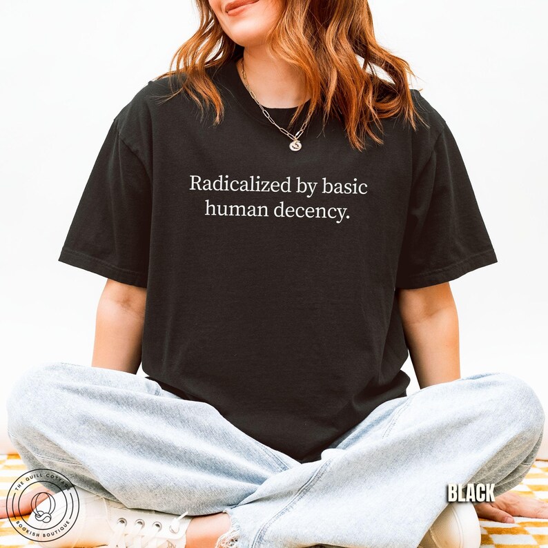 May include: Black t-shirt with the text "Radicalized by basic human decency" in white. The shirt is a crew neck style and appears to be made of a soft material. The word "BLACK" is printed in white at the bottom right.