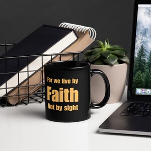 Puede incluir: Taza de café negra con una inscripción blanca que dice "For we live by Faith Not by sight". El texto está en color dorado.