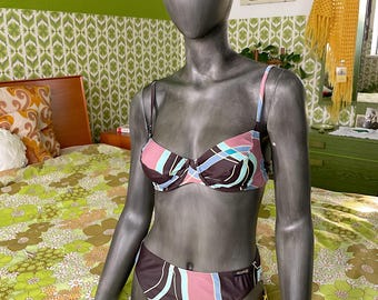 May include: A two-piece bikini set on a mannequin. The bikini top and bottom feature a brown, pink, and teal geometric pattern. The straps are light blue. The mannequin is gray and stands on a floral patterned bedspread.