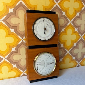 Könnte beinhalten: Eine rechteckige Wetterstation aus Holz mit zwei übereinander angeordneten runden Anzeigen. Die obere Anzeige ist ein Barometer mit weißem Zifferblatt und schwarzen Zeigern. Die untere Anzeige ist ein Raumklimamesser mit den Beschriftungen RAUM-KLIMAMESSER, FEUCHTE und CELSIUS, schwarzen Zeigern und goldfarbenen Rändern.