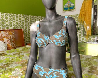 May include: A two-piece bikini set displayed on a mannequin. The bikini features a brown base color with a blue floral pattern. The top is a bra-style with adjustable straps, and the bottoms are a classic cut. The mannequin is positioned on a floral patterned bedspread.