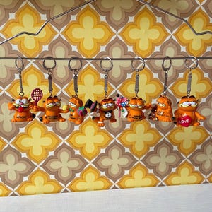 Könnte beinhalten: Eine Sammlung von Garfield-Schlüsselanhängern, die jeweils die orangefarbene Katze in verschiedenen Posen und Outfits zeigen. Einige Schlüsselanhänger haben Accessoires wie einen Tennisschläger, einen Zylinder oder ein Herz mit dem Wort "LOVE". Sie hängen an einem Metallbügel vor einer Retro-Blumentapete.