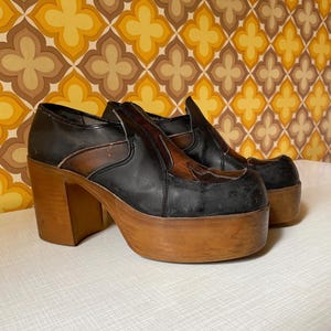 May include: A pair of vintage platform shoes. The shoes have a black leather upper with brown accents and a thick wooden platform sole. The heels are also made of wood. The shoes are in good condition and appear to be from the 1970s.