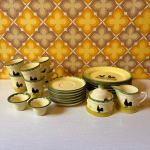 Könnte beinhalten: Ein Vintage-Keramik-Geschirrset mit einem Hahn-Motiv. Das Set umfasst Tassen, Untertassen, Schalen, Teller, eine Zuckerdose und einen Sahnekännchen. Die Teile sind cremefarben mit grünen Verzierungen und Hahn-Illustrationen.