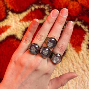 Könnte beinhalten: Fünf messingfarbene Ringe mit klaren, runden Glaskabochons, die ein dunkles, abstraktes Design zeigen. Die Ringe werden an einer Hand getragen. Der Hintergrund ist ein strukturierter, rot-oranger Teppich.