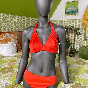 May include: A bright orange bikini set on a mannequin. The two-piece swimsuit has a halter neck top and matching low-rise bottoms. The mannequin is placed on a floral patterned bedspread, highlighting the swimwear's colour and design.