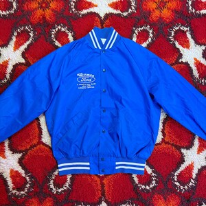 Könnte beinhalten: Eine leuchtend blaue Vintage-Jacke mit weißen Streifen an Kragen, Bündchen und Bund. Die Jacke zeigt das Logo und den Text "Wittman Ford" auf der Brust. Die Jacke hat einen Knopfverschluss und einen klassischen Bomberstil.