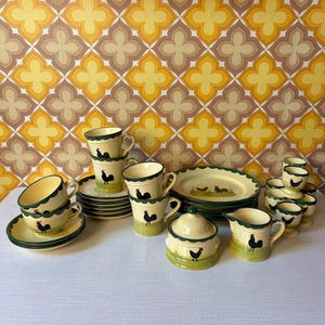 Może przedstawiać: Zestaw do herbaty w stylu vintage, wykonany z kremowej ceramiki z zielonymi obramowaniami i wzorem koguta. Zestaw zawiera filiżanki, spodki, talerze, cukiernicę i dzbanek do śmietanki. Tło to retro tapeta w żółte i brązowe kwiaty.