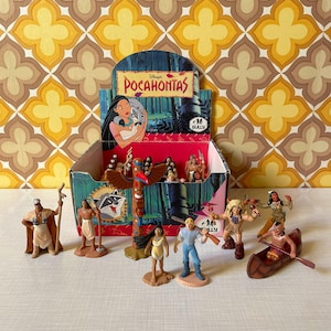 May include: A collection of Disney's Pocahontas figurines, including characters and a totem pole, displayed in a themed box. The box features the Pocahontas logo and character art. The background has a retro floral pattern.