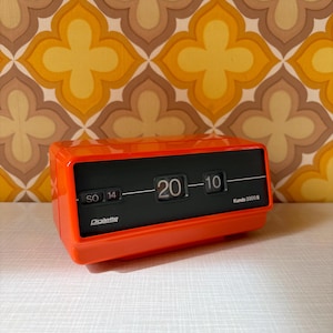 Puede incluir: Reloj de solapa naranja Digiletta Kundo 2000 S que muestra la hora 20:10 sobre un fondo negro. El día es sábado, 14. El reloj está sobre un papel pintado floral retro.