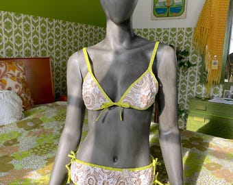 May include: A two-piece bikini set displayed on a mannequin. The bikini features a white lace design with yellow trim and ties. The background includes a floral patterned bedspread and a green wall.