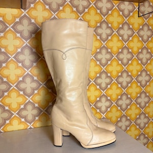 Puede incluir: Un par de botas altas color crema con tacón de bloque. Las botas tienen una punta redondeada y un detalle de costura decorativa en la parte superior. Las botas están hechas de un material liso y brillante.