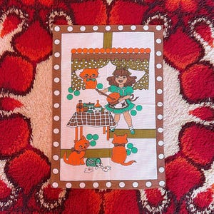 May include: Vintage decorative kitchen towel featuring a whimsical illustration. The design includes a girl playing a stringed instrument, orange cats, and a table setting. The towel has a brown border with white polka dots and a white background.