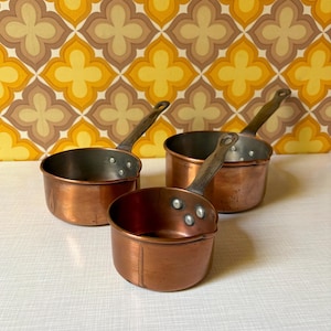 Könnte beinhalten: Drei Kupfertöpfe mit Metallgriffen. Die Pfannen sind unterschiedlich groß und stehen auf einer weißen Oberfläche. Der Hintergrund zeigt ein Retro-Blumenmuster in Gelb-, Braun- und Weißtönen.