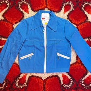 May include: A vibrant blue jacket with a white zip and two front pockets. The jacket features a collar and a tag. Set against a red and orange patterned rug.