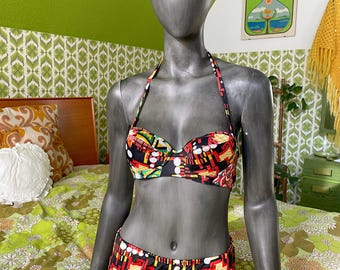 May include: A two-piece bikini set with a vibrant geometric pattern in red, yellow, green, and black. The halter-style top and high-waisted bottoms are displayed on a mannequin. The background features a floral bedspread and a green wall.