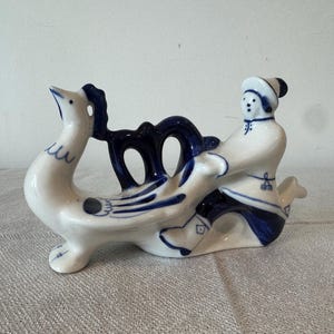 Puede incluir: Una figura de cerámica blanca y azul que representa a una persona montada en un cisne. El cisne tiene detalles azules, y el jinete está vestido de blanco con detalles azules. La figura es un objeto decorativo.