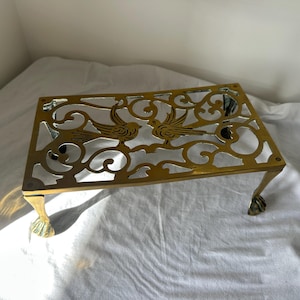 May include: A rectangular brass trivet featuring an intricate openwork design of two birds facing each other amidst swirling scrollwork patterns. The piece is supported by four ornate legs that terminate in detailed claw and ball feet.