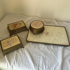 Może przedstawiać: Kolekcja czterech przedmiotów toaletowych w stylu vintage, obejmująca dwa prostokątne metalowe pudełka, okrągły metalowy pojemnik oraz prostokątną tacę. Każdy element posiada ozdobną ramę w kolorze złota oraz środkowy panel z kwiatowym haftem.