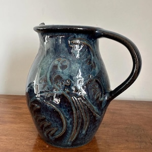 Può includere: Brocca in ceramica fatta a mano con smalto blu scuro e intricati motivi floreali in rilievo. La brocca presenta un manico grande e curvo e un beccuccio leggermente svasato. L'altezza totale è di circa 25 cm.