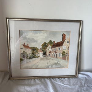 May include: A framed watercolor painting depicting a quiet village street scene with brick buildings, a pub sign, and trees under a cloudy sky. The artwork is enclosed in a thin, metallic silver frame with a white mat border.