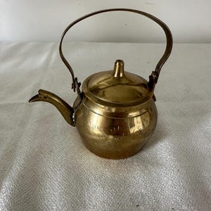 Może przedstawiać: Mały, zabytkowy mosiężny czajniczek z zakrzywionym uchwytem i dzióbkiem. Czajniczek posiada pokrywkę i jest zdobiony wytrawionymi liniami. Mosiądz ma ciepły, złoty kolor. Czajniczek jest prawdopodobnie używany do parzenia herbaty.