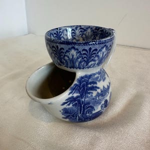 Op de afbeelding: Een tweedelige scheerketel van blauw en wit porselein. De bovenste kom heeft een handvat en een gedetailleerd bloemmotief. Het onderste gedeelte heeft een landschapsscène en een tuit voor water.