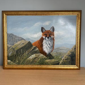 May include: A framed oil painting of a fox in a natural landscape. The fox has reddish-brown fur and a white chest, set against a backdrop of rocks, grass, and mountains under a cloudy sky. The frame is gold.