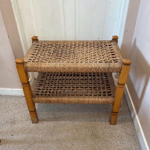 May include: A two-tiered, light brown woven bench with a rectangular shape. The bench has a woven top and bottom shelf, supported by four wooden legs. The legs are a lighter shade of brown and have a square shape.