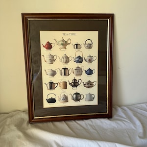 May include: A framed wall art print titled TEA-TIME featuring a grid of nineteen various teapots in diverse colours, shapes, and materials including ceramic, metal, and porcelain, set against a cream background with a dark brown mat and wood frame.